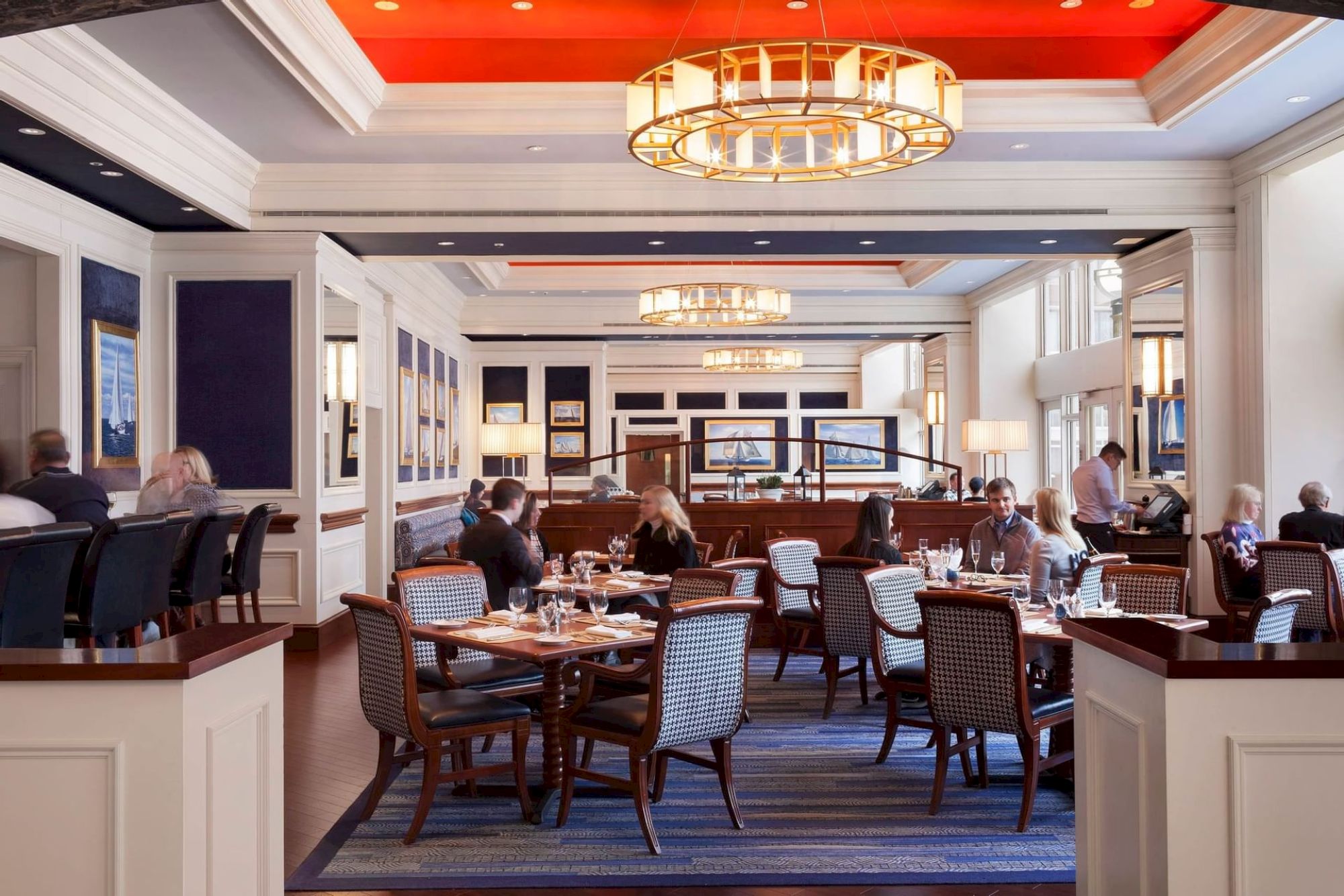 A stylish restaurant interior with people dining, featuring elegant lighting, wooden furniture, and a vibrant red ceiling accent.