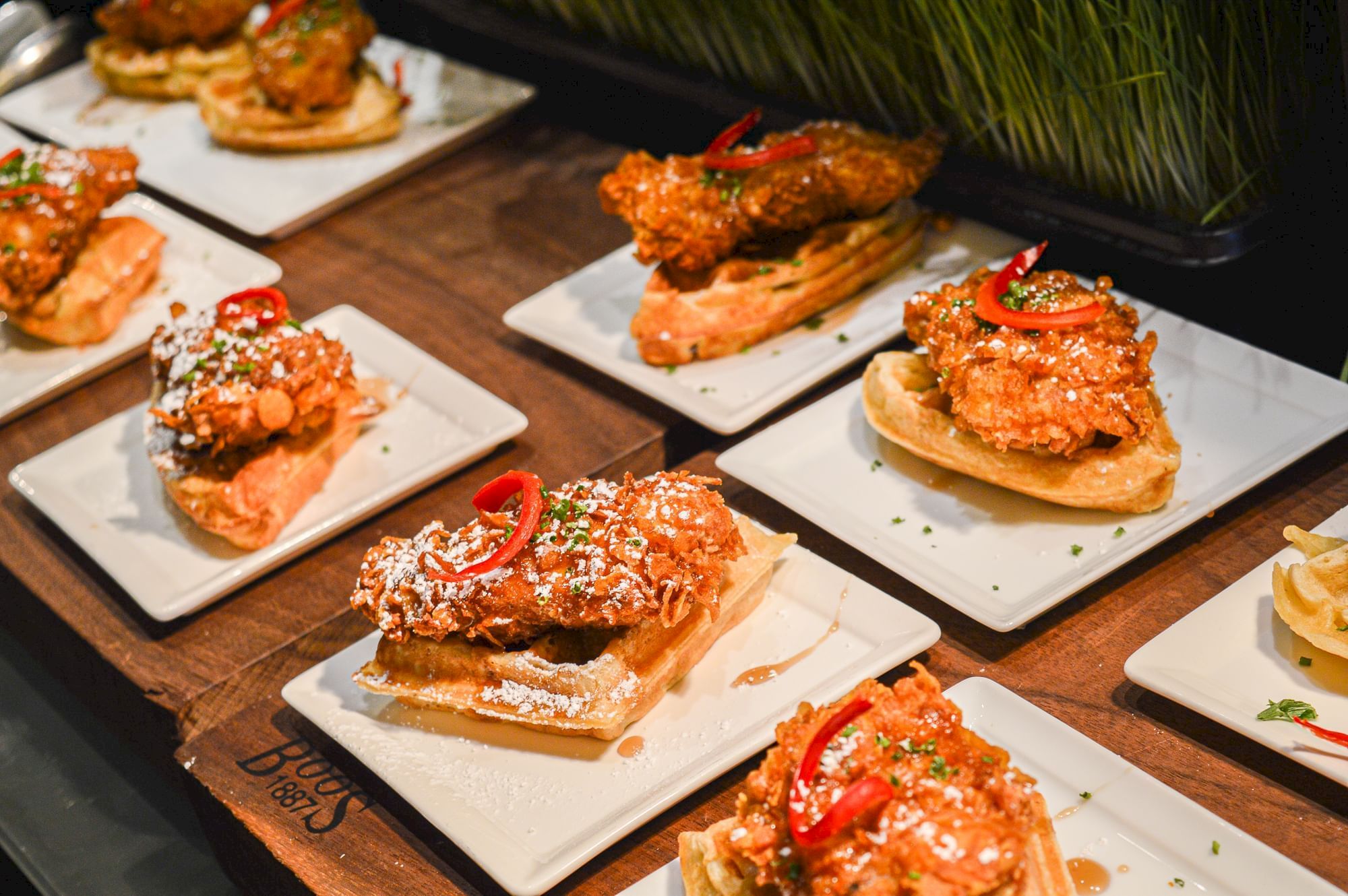 The image shows plates of fried chicken on waffles, garnished with sliced red peppers and herbs, arranged on a wooden board.