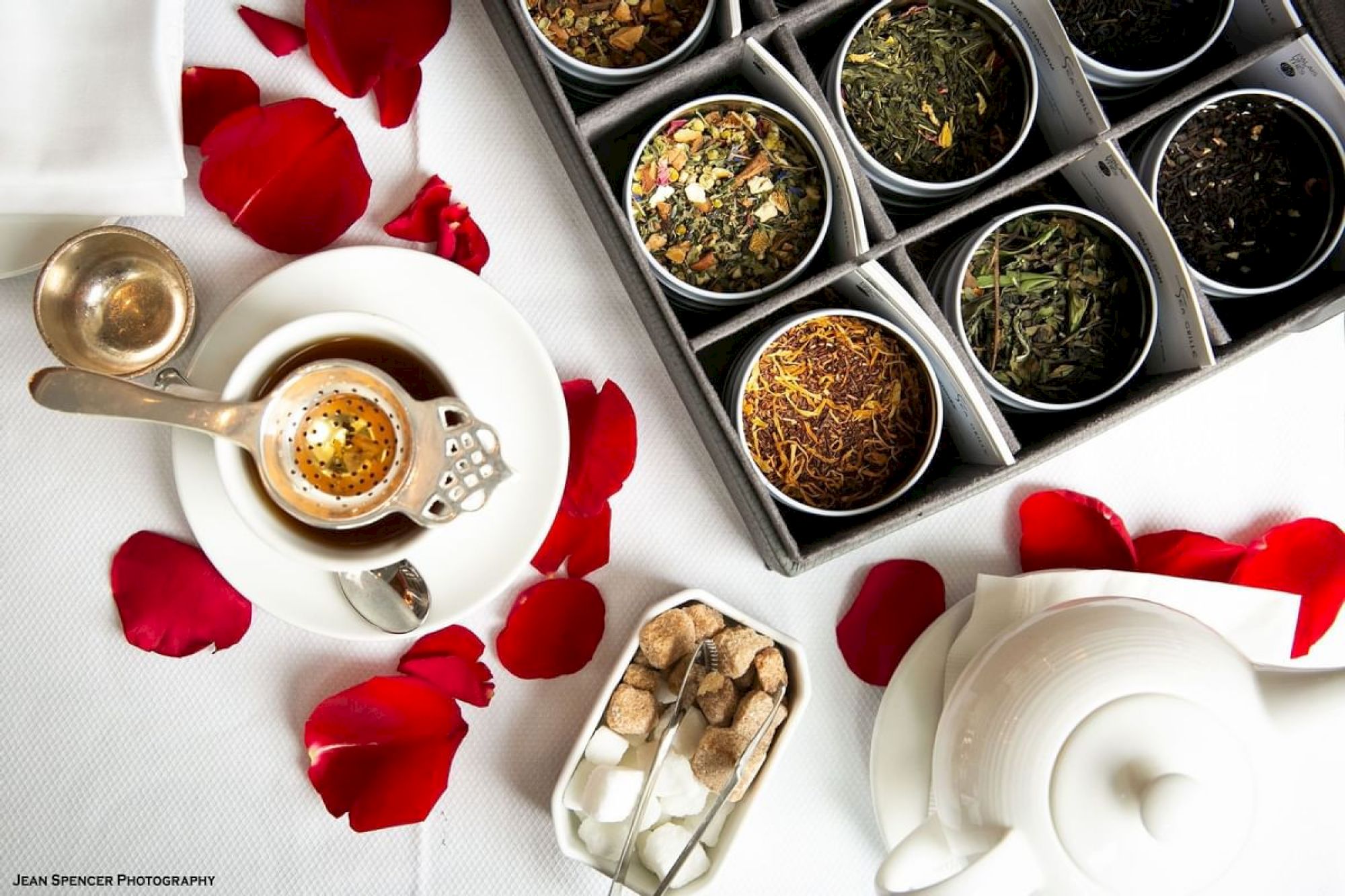 A tea set with various loose leaf teas, a teapot, and sugar cubes on a white tablecloth decorated with red rose petals.