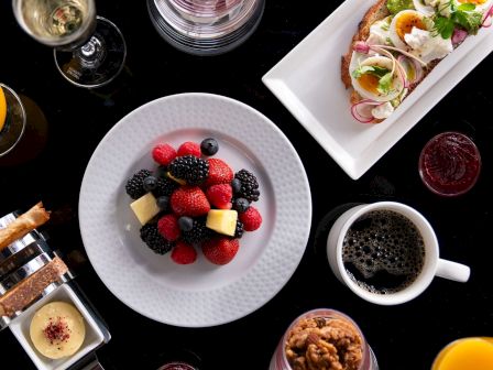 Aerial view of a breakfast spread with fruit bowl, toast, drinks, pastries, and coffee on a dark table.