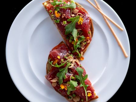 Two gourmet open-faced sandwiches topped with tuna, greens, and garnishes on a white plate, with two breadsticks beside them.