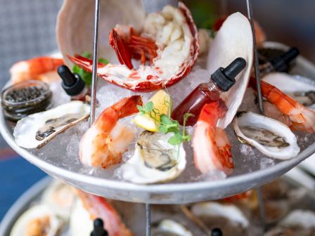 A seafood platter featuring shrimp, oysters, and lobster on ice, decorated with lemon slices and garnished with small sauce bottles for flavor.