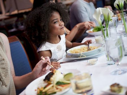 A young girl enjoys pasta at a table with others, surrounded by plates of food and flowers.