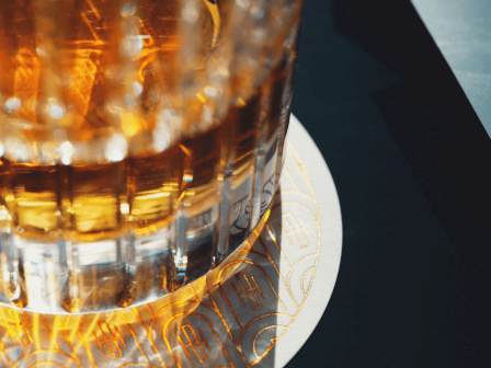 A glass of amber liquid, likely whiskey, on a patterned coaster. The scene is dimly lit, creating a warm, inviting atmosphere.