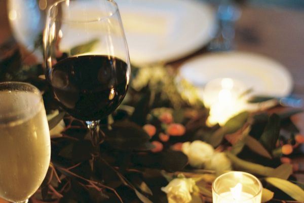 A table with a wine glass, beer, greenery, a lit candle, and plates, set for a cozy dinner.