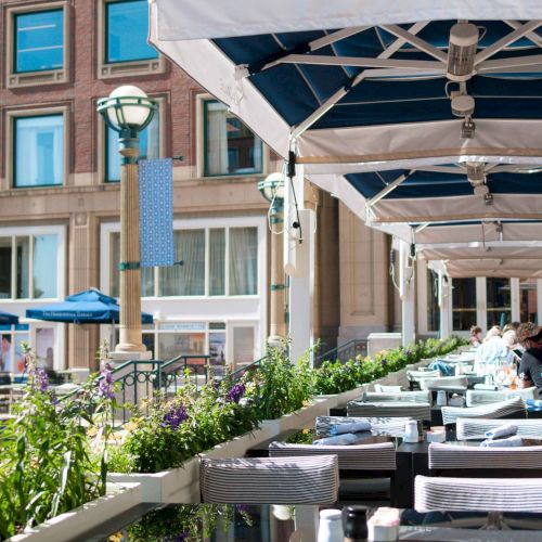 The image shows an outdoor restaurant seating area with blue umbrellas, tables set with white linens, and a building in the background.