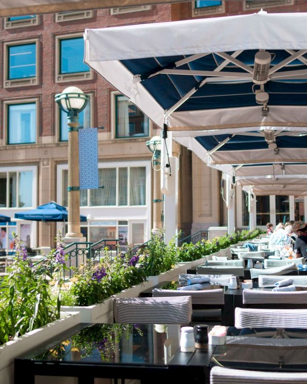 The image shows an outdoor restaurant seating area with blue umbrellas, tables set with white linens, and a building in the background.