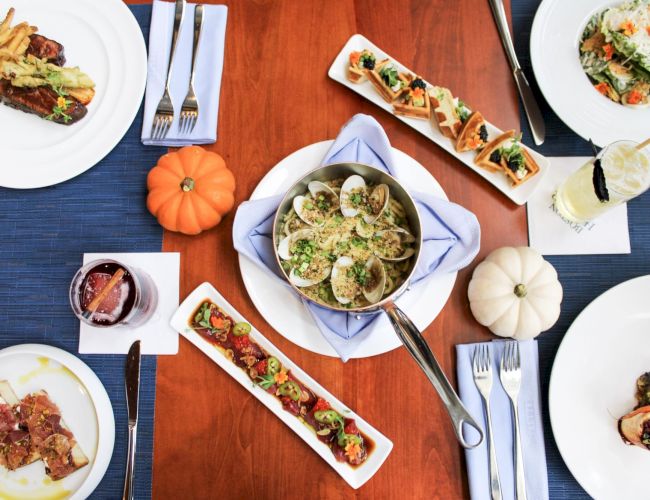 The image shows a table with various dishes, drinks, and decorative mini pumpkins set for a meal on wooden surface with blue placemats.