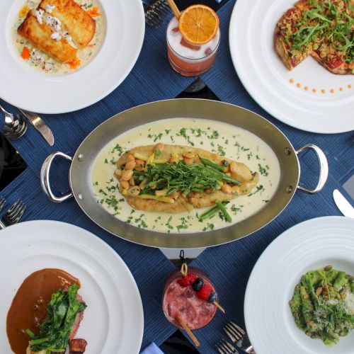 A dining table with various dishes: pasta, meat, pizza, sauces, and drinks, all beautifully arranged on a blue tablecloth.