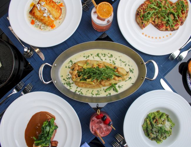 A dining table with various dishes: pasta, meat, pizza, sauces, and drinks, all beautifully arranged on a blue tablecloth.