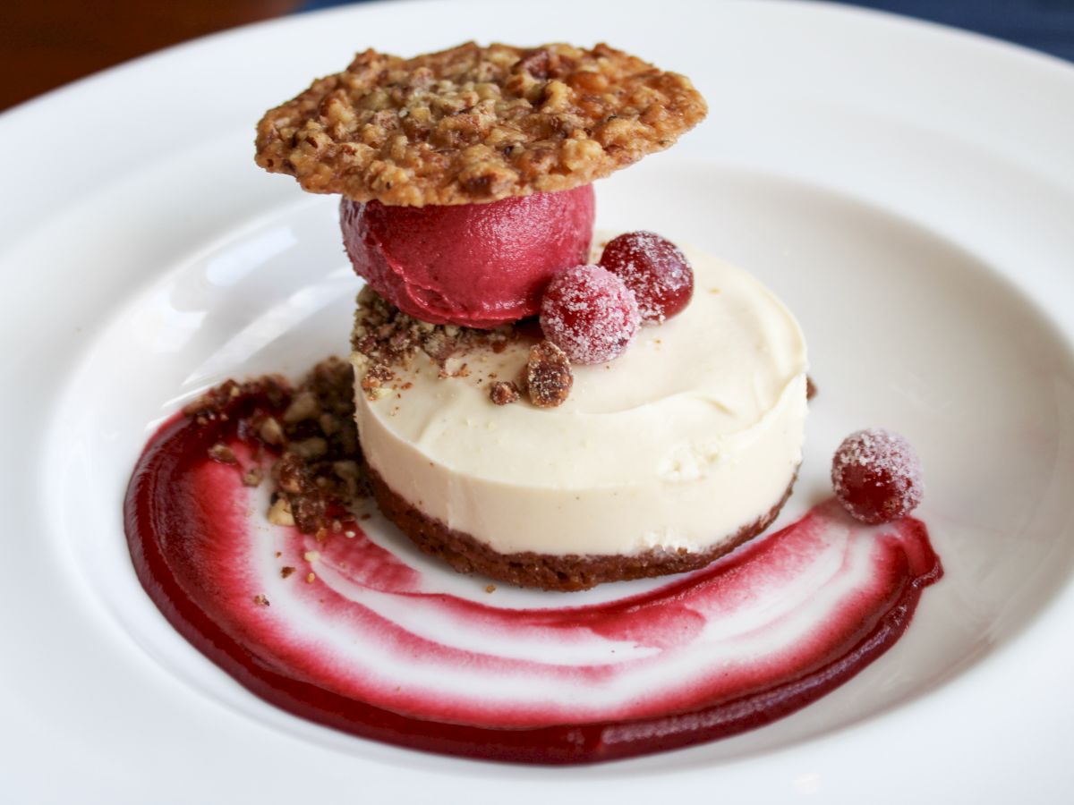 A dessert with a cream base, topped with red sorbet, a cookie, and garnished with berries and a red sauce swirl on the plate.