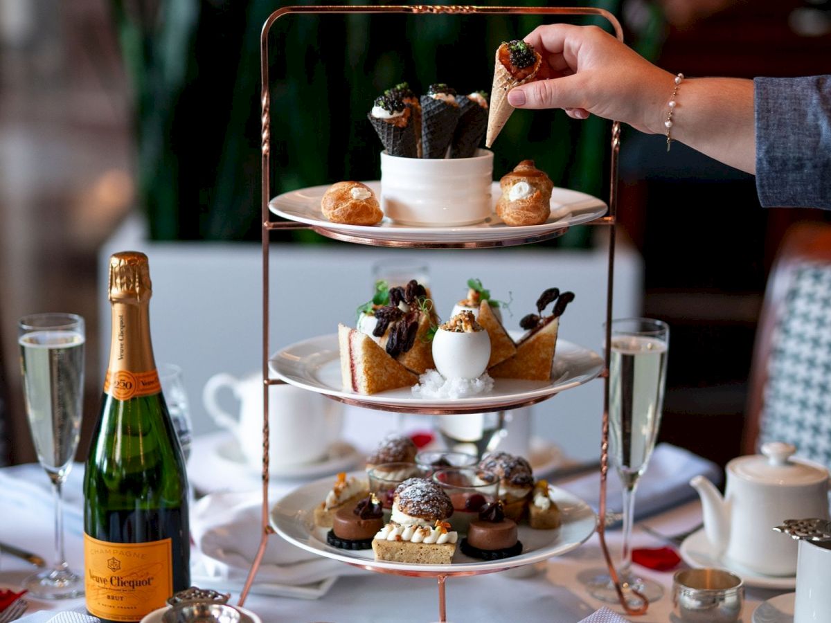 A tiered tray with assorted pastries and desserts, champagne, teapots, and cups on a table set for afternoon tea.