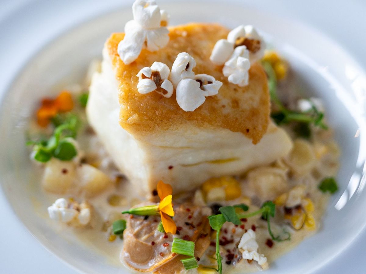 A gourmet dish with a piece of seared fish, garnished with popcorn, microgreens, and edible flowers on a colorful cream-based sauce.