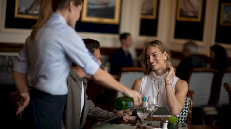 A server pouring wine for a couple at a restaurant. The setting is elegant with framed artwork on the walls in the background.