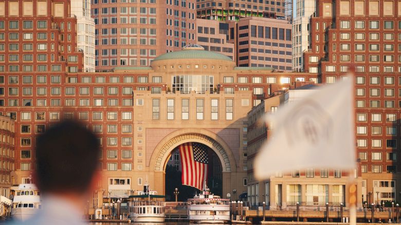 The image shows a patriotic scene with a large American flag hanging in a building archway, surrounded by cityscape and waterfront views.