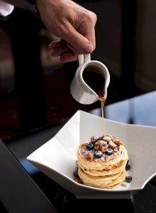 A person pouring syrup over a stack of pancakes topped with blueberries and nuts, placed in a white bowl.