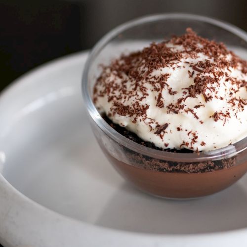 A chocolate dessert in a glass dish with whipped cream and chocolate shavings on top, placed on a white plate.