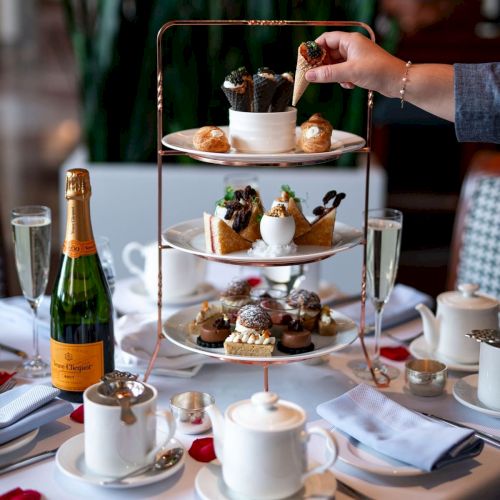 A three-tiered afternoon tea setup with assorted pastries, champagne, tea, and scattered rose petals on a table.