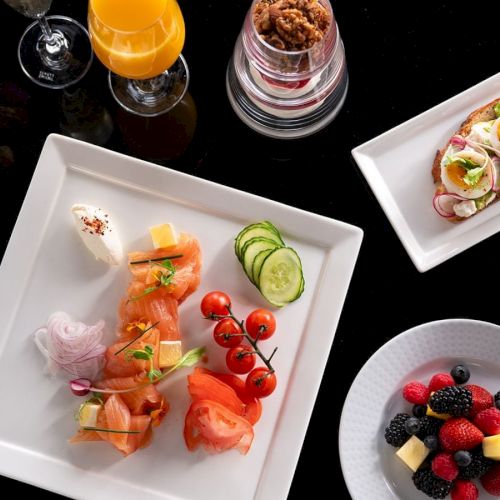 A breakfast spread featuring smoked salmon, toast, fruit, coffee, juice, and a yogurt or parfait with granola on a black surface.