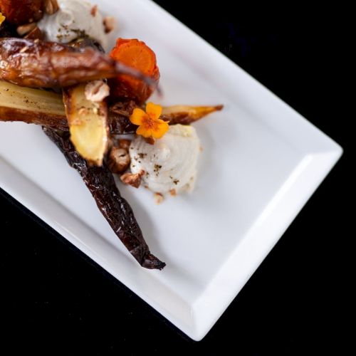 The image shows a gourmet dish with assorted roasted vegetables, nuts, and garnishes on a rectangular white plate.