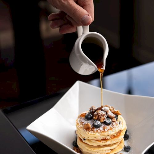 A person is pouring syrup over a stack of pancakes topped with blueberries and nuts, on a white plate.