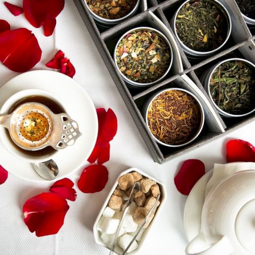 The image shows an assortment of tea leaves, a teapot, a cup of tea, sugar cubes, and rose petals on a white tablecloth.