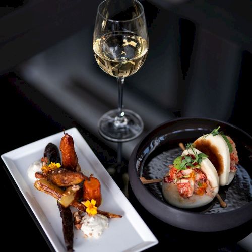 The image shows a glass of white wine, a plate with cooked vegetables, and another with bao buns featuring a garnish on a glossy surface.