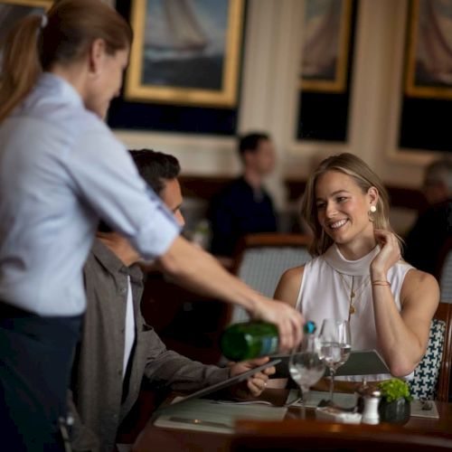 A server pours water for dining guests, as a woman smiles at a restaurant with nautical-themed decor.