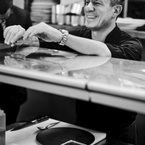 A person smiling while preparing food in a kitchen, with a workspace featuring knives and ingredients.