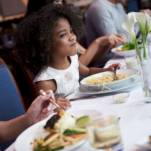 A family is enjoying a meal at a restaurant. A girl is eating pasta, surrounded by various dishes and glasses on a white tablecloth.