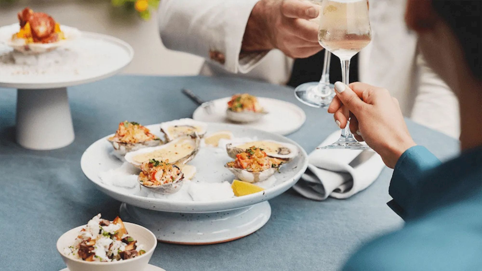 A table setting with oysters, a small salad, a glass of white wine, and a person in a suit reaching for it.