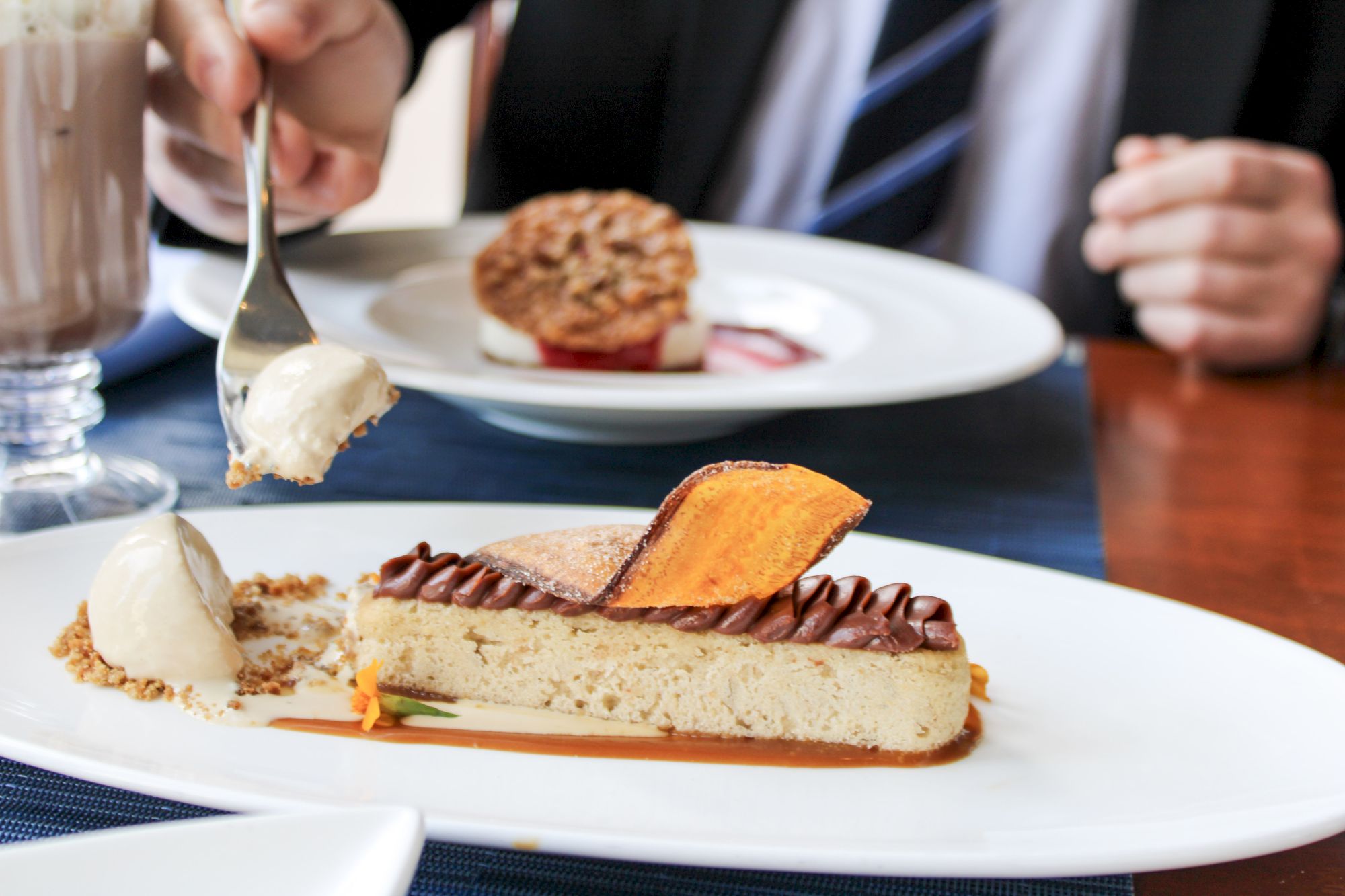 A slice of dessert with cream and a crispy garnish is being enjoyed. Another dessert and a drink are in the background on the table.