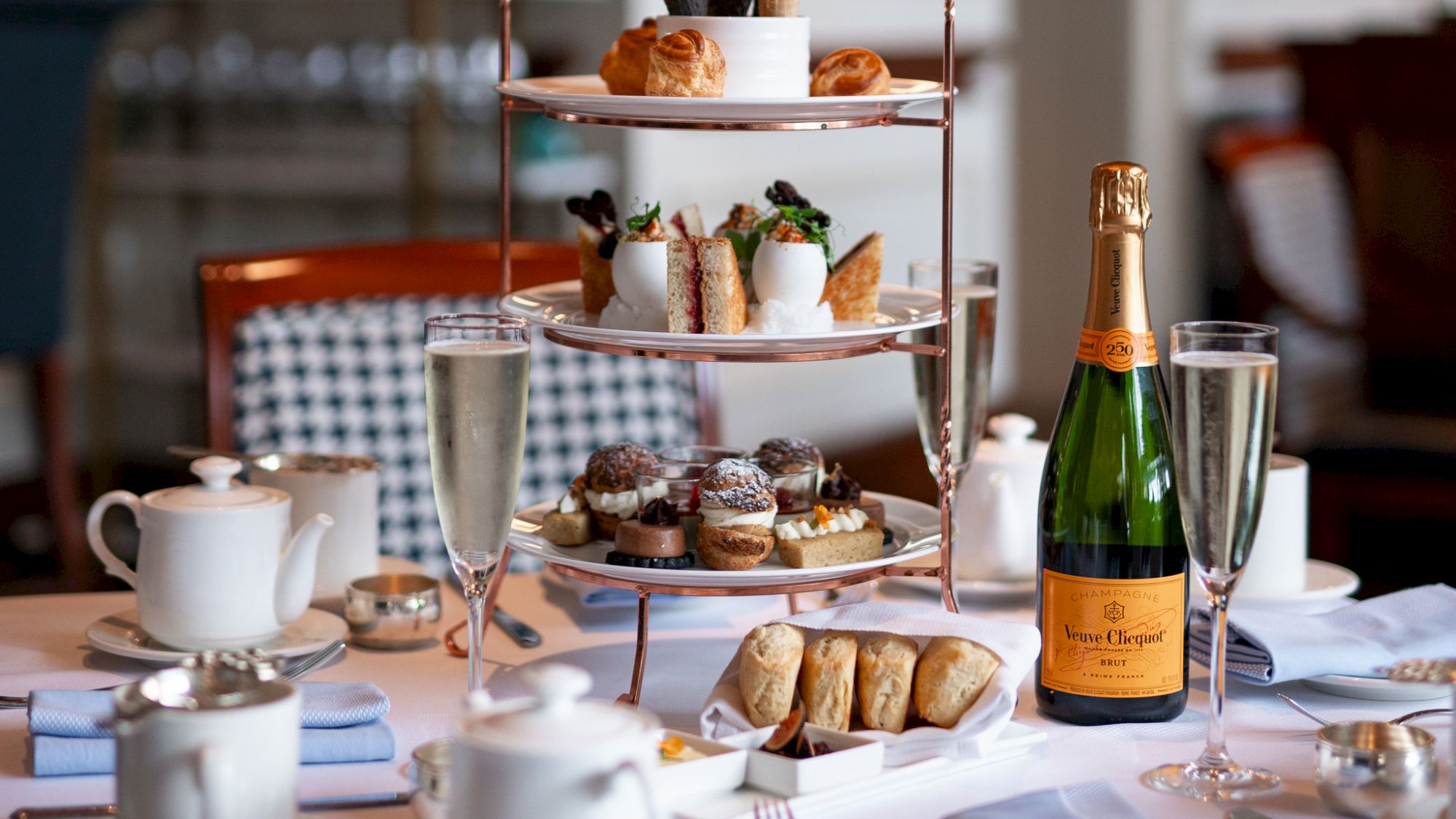 An elegant afternoon tea setup with a three-tiered stand, pastries, sandwiches, champagne bottle, and glasses on a table.