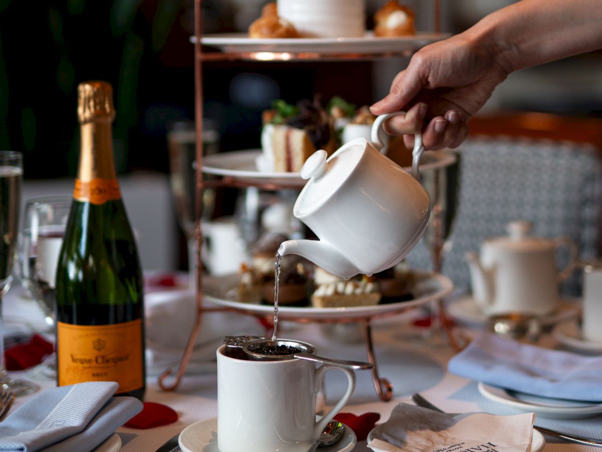 A person pours tea into a cup at a table set with afternoon tea, featuring a champagne bottle, rose petals, and tiered pastries.