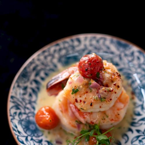 A gourmet dish featuring shrimp, cherry tomatoes, and herbs on a decorative blue and white plate, presented with a light sauce.