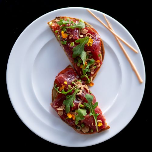 Two tostadas topped with tuna, arugula, and garnishes on a white plate with two thin breadsticks.